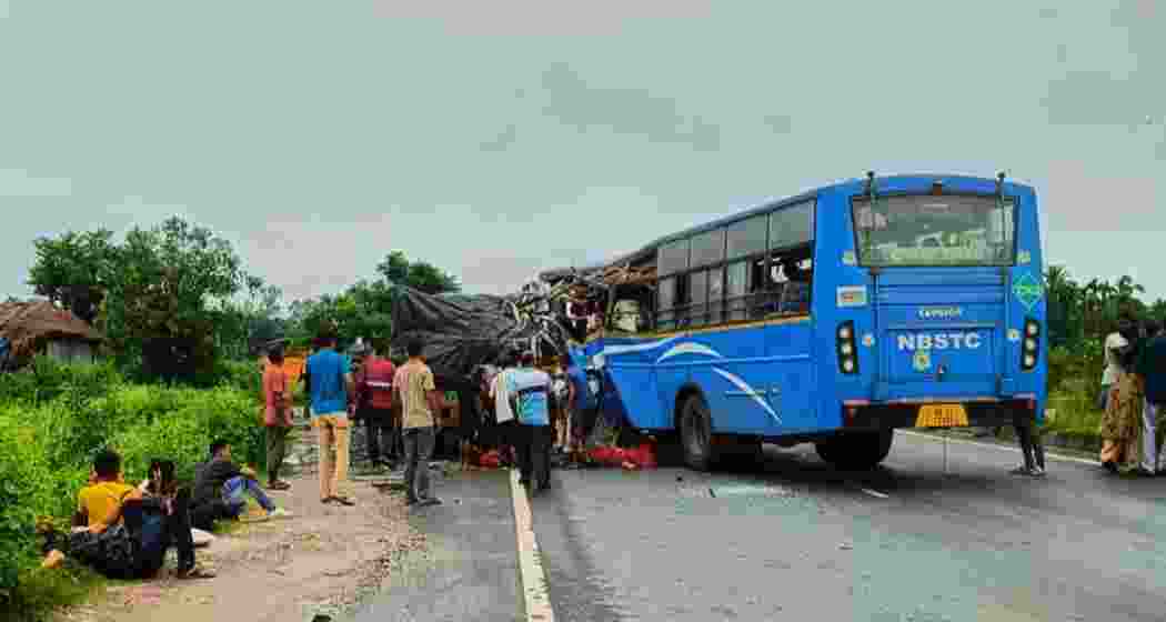 Local residents gather around the damaged NBSTC bus after it rammed into a stationary trailer on NH-27 in Jalpaiguri district on Sunday, while injured passengers await assistance by the roadside.